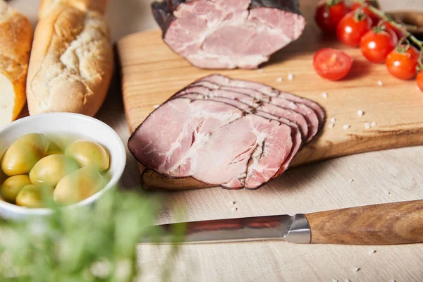 Selective focus of tasty ham on cutting board with knife, cherry tomatoes, olives and baguette on wooden table — Stock Photo