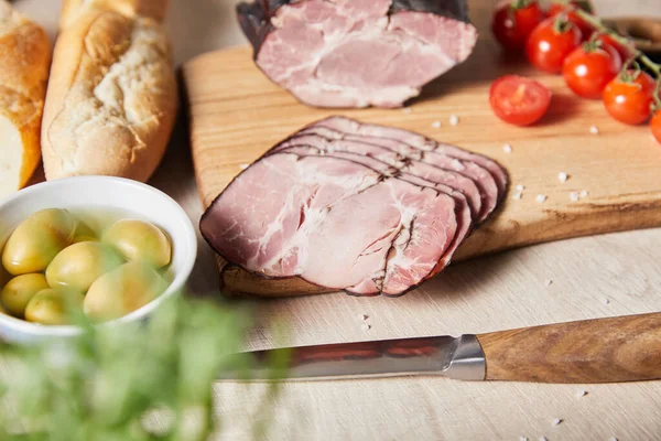 Foyer sélectif de jambon savoureux sur planche à découper avec couteau, tomates cerises, olives et baguette sur table en bois — Photo de stock