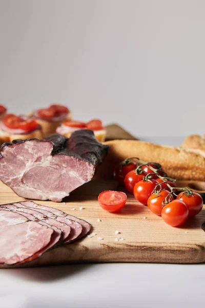 Selettiva messa a fuoco del gustoso prosciutto sul tagliere con pomodorini e baguette vicino alla tartina isolata sul grigio — Foto stock