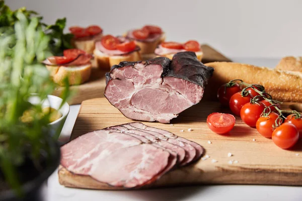 Foco seletivo de presunto saboroso na tábua de corte com tomate cereja e baguete perto de canapé isolado em cinza — Fotografia de Stock