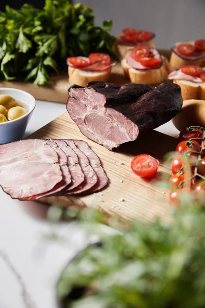 Selective focus of tasty ham on cutting board with parsley, cherry tomatoes, olives and baguette near canape — Stock Photo