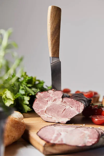 Selective focus of knife in tasty ham on cutting board with parsley, cherry tomatoes and baguette isolated on grey — Stock Photo