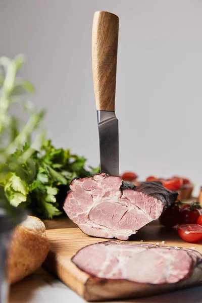 Selektiver Schwerpunkt des Messers in schmackhaftem Schinken auf Schneidebrett mit Petersilie, Kirschtomaten und Baguette isoliert auf grau — Stockfoto