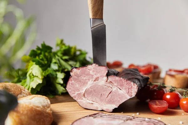 Enfoque selectivo de cuchillo en sabroso jamón en tabla de cortar con perejil, tomates cherry y baguette aislado en gris - foto de stock