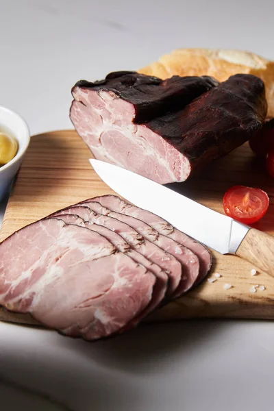 Enfoque selectivo de sabroso jamón en la tabla de cortar con cuchillo, tomate cherry y baguette en la superficie blanca - foto de stock