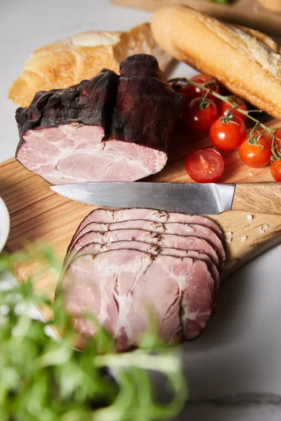 Enfoque selectivo de sabroso jamón en la tabla de cortar con cuchillo, tomates cherry y baguette - foto de stock