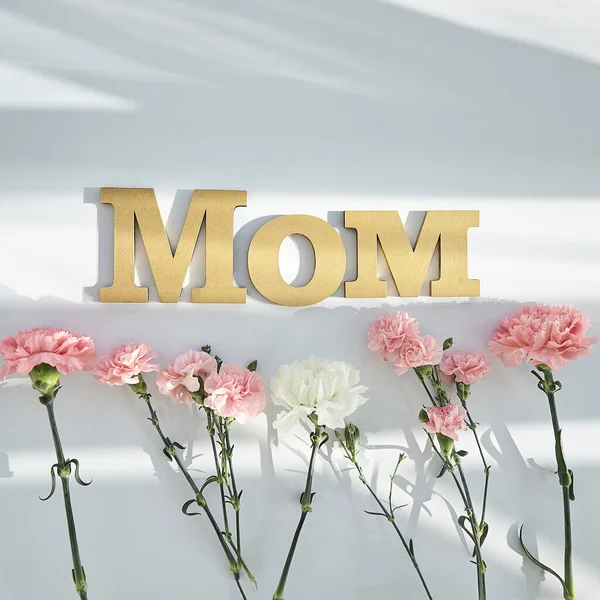 Top view of pink and white carnations and mom lettering on white background with sunlight and shadows — Stock Photo