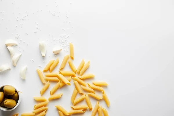 Vista dall'alto di pasta, aglio, ciotola con olive e sale marino su fondo bianco — Foto stock