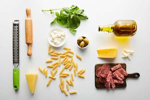 Flat lay with meat platter, bottle of olive oil, rolling pin, grater, pasta and ingredients on white — Stock Photo