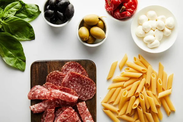 Vue du dessus du plateau de viande, des pâtes, des feuilles de basilic et des bols avec des ingrédients sur fond blanc — Photo de stock