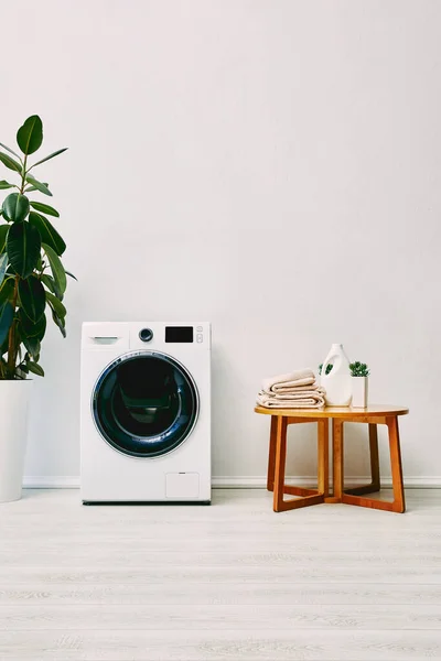 Planta verde perto da máquina de lavar roupa e mesa de café de madeira com toalhas e garrafa de detergente no banheiro — Fotografia de Stock