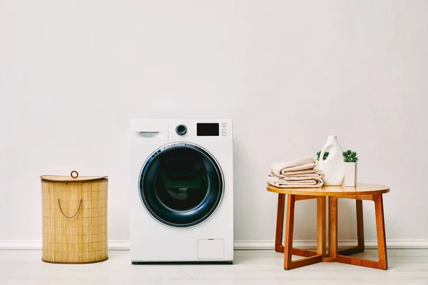 Machine à laver moderne près du panier à linge, table basse avec serviettes, bouteille de détergent et plante dans la salle de bain — Photo de stock
