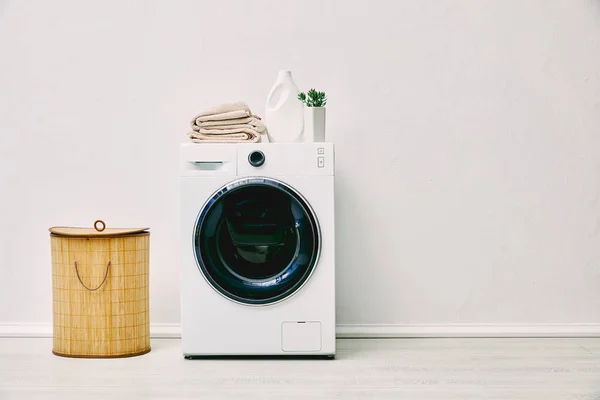 Бутылка моющего средства, полотенца и растение на стиральной машине возле корзины для белья в ванной комнате — стоковое фото