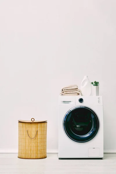 Бутылка моющего средства, полотенца и зеленые растения на современной стиральной машине возле корзины для белья в ванной комнате — стоковое фото