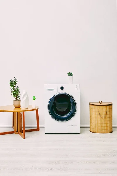 Plantas verdes e garrafas perto da mesa de café, cesta de roupa e máquina de lavar roupa moderna no banheiro — Fotografia de Stock