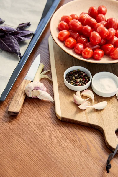 Vista de ángulo alto de la bandeja del horno, placa y tabla de cortar con ingredientes cerca del cuchillo sobre fondo de madera - foto de stock