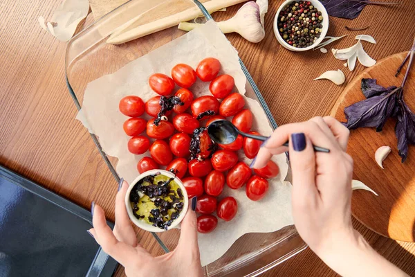 Vista ritagliata della donna che aggiunge foglie di basilico tagliate con olio d'oliva ai pomodori in teglia su fondo di legno — Foto stock