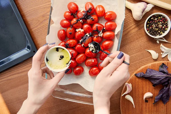 Vista recortada de la mujer preparando tomates cerca de la bandeja del horno y tabla de cortar con ingredientes sobre fondo de madera - foto de stock