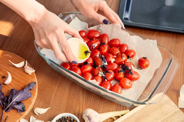 Vista recortada de la mujer añadiendo aceite de oliva a los tomates con ingredientes cerca de la bandeja del horno y tablas de cortar sobre fondo de madera - foto de stock