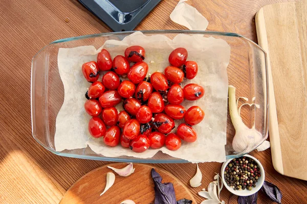 Vista superior de tomates con hojas de albahaca cortadas sobre papel de hornear cerca de tablas de cortar, ajo y tazón con pimienta sobre fondo de madera - foto de stock