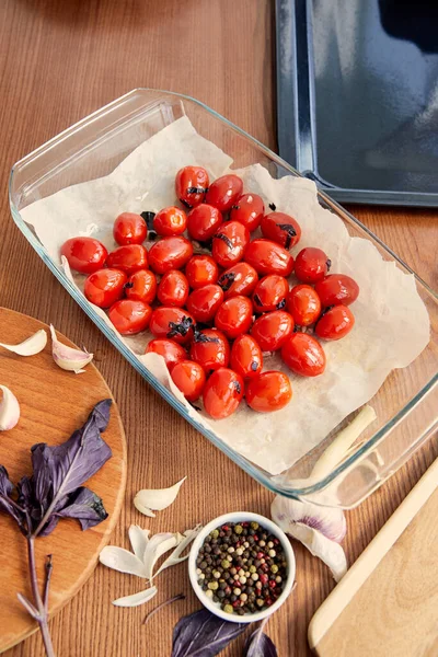 Vista de ángulo alto de la bandeja para hornear con tomates cerca de la bandeja del horno, tablas de cortar con ajo, hojas de albahaca y tazón con pimienta sobre fondo de madera - foto de stock