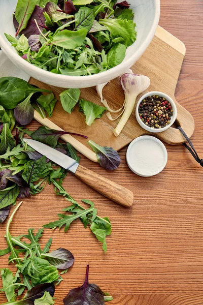 Top view of bowls with pepper, salt and salad ingredients on cutting board with knife on wooden background — Stock Photo