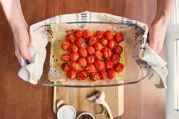 Visão superior da mulher com guardanapo segurando assadeira com tomates cozidos perto de alho e tigelas com sal e pimenta na tábua de corte — Fotografia de Stock