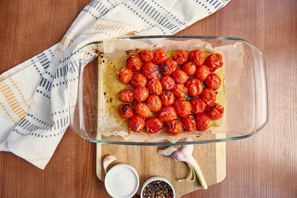 Top view of baking dish with cooked tomatoes near garlic and bowls with salt and pepper on cutting board near napkin on wooden background — Stock Photo