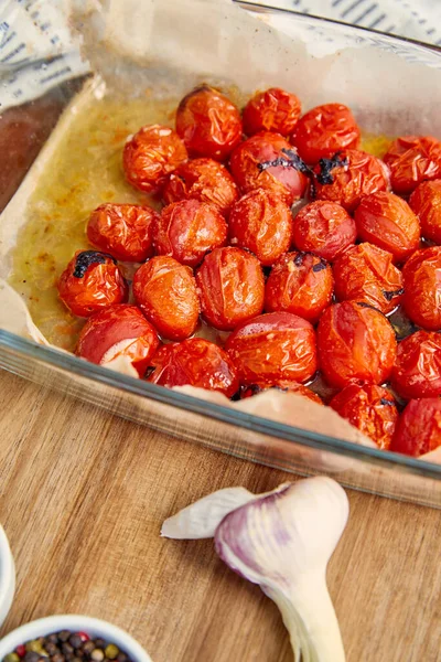 High angle view of prepared tomatoes in baking dish near garlic on wooden background — Stock Photo