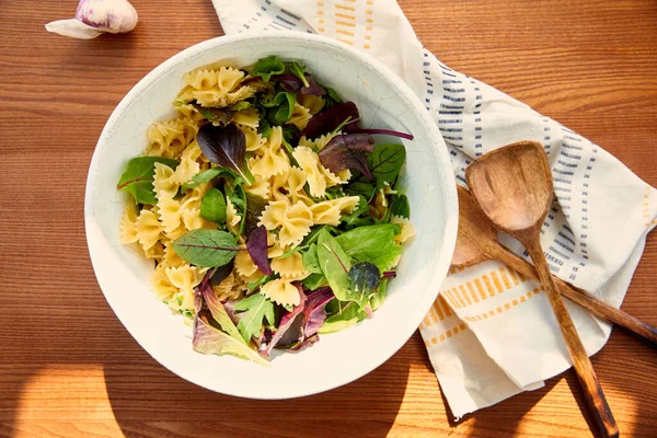 Top view of bowl with pasta salad ingredients near garlic, napkin and spatulas on wooden background — Stock Photo