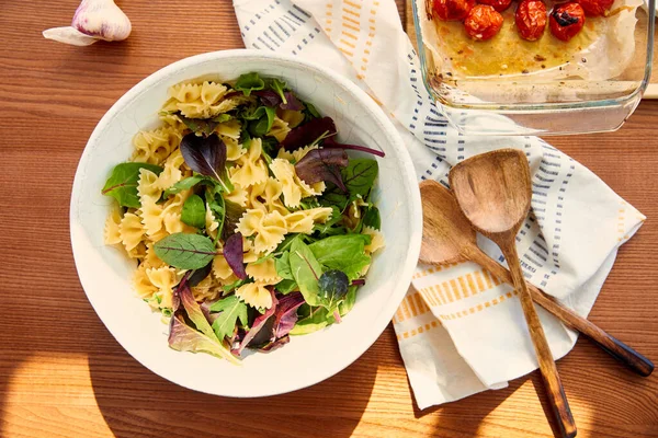 Vista superior da tigela com ingredientes de salada de macarrão perto de alho, guardanapo, espátulas e assadeira com tomates em fundo de madeira — Fotografia de Stock