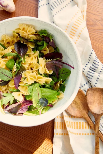 Top view of bowl with pasta salad ingredients with spatulas, napkin and garlic on wooden background — Stock Photo