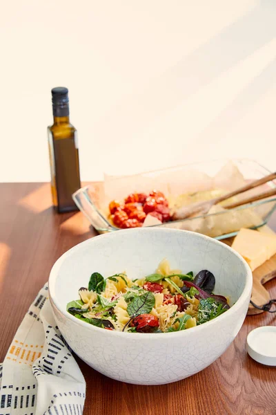 Bol avec salade de pâtes et sel près d'une bouteille d'huile d'olive, plat de cuisson et serviette sur fond bois et beige — Photo de stock