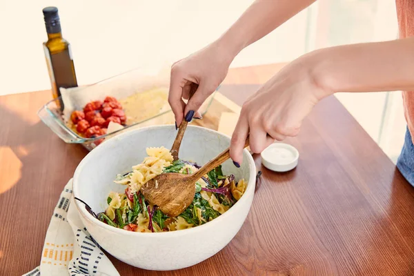 Cropped view of woman mixing pasta salad in bowl with spatulas near napkin at table in kitchen — Stock Photo