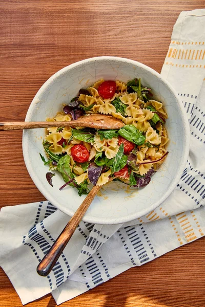 Vista superior del tazón con ensalada de pasta y espátulas cerca de servilleta sobre fondo de madera - foto de stock