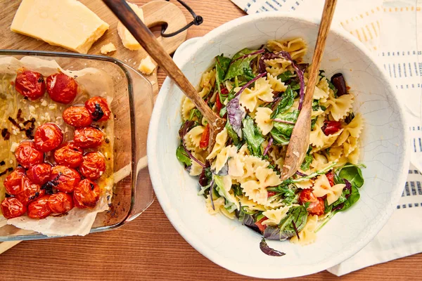 Top view of bowl with pasta salad and spatulas near ingredients on cutting board and napkin on wooden background — Stock Photo