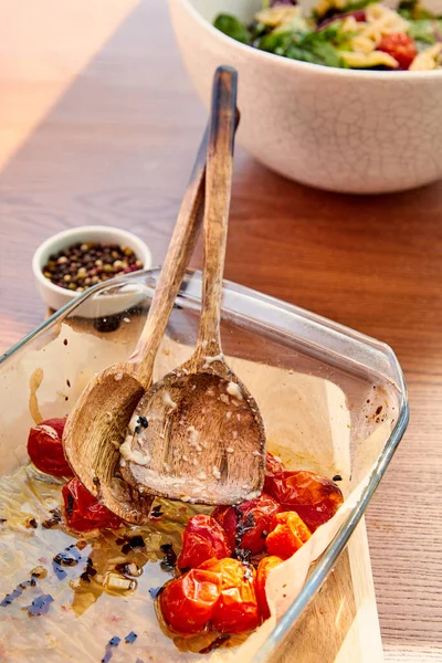 High angle view of baking dish with spatulas and tomatoes on cutting board near bowls  with pepper and pasta salad on wooden background — Stock Photo