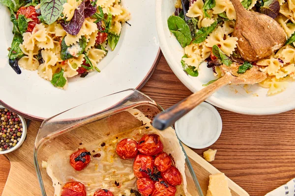 Draufsicht auf Teller und Schüssel mit Nudelsalat und Spateln in der Nähe von Schneidebrett und Backform mit Zutaten auf Holzgrund — Stockfoto