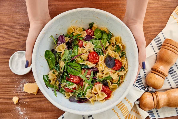 Vista recortada de la mujer sosteniendo tazón con ensalada de pasta cerca de parmesano y servilleta con pimienta y molinos de sal sobre fondo de madera - foto de stock