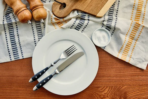 Vue du dessus de la plaque avec fourchette et couteau près de la serviette avec planche à découper, moulins à poivre et sel sur fond en bois — Photo de stock