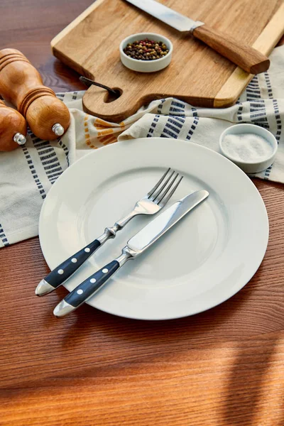 Vista ad alto angolo del piatto con posate vicino tovagliolo, tagliere, ciotole, pepe e sale mulini su sfondo di legno — Foto stock