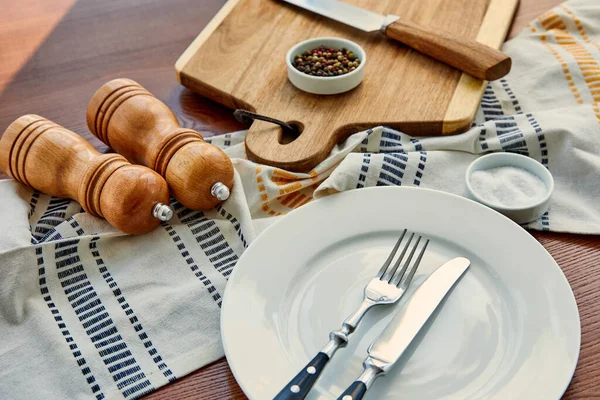 High angle view of plate with cutlery near cloth, cutting board, bowls, pepper and salt mills on wooden background — Stock Photo