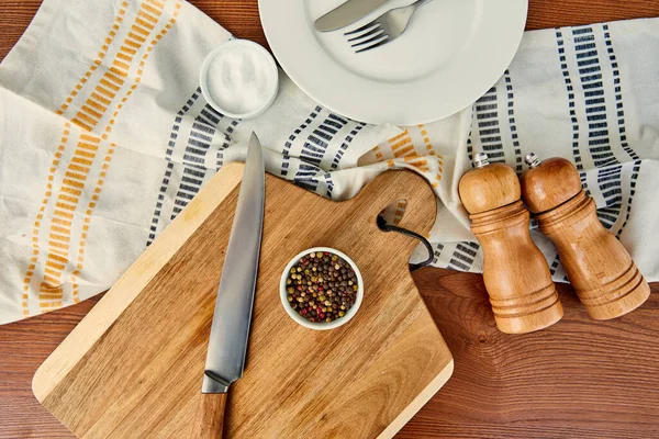 Top view of plate with cutlery near cloth, cutting board, bowls, pepper and salt mills on wooden background — Stock Photo