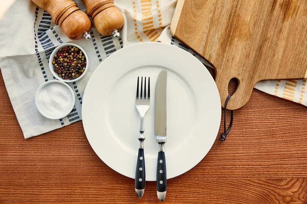 Вид сверху тарелки с ножом и вилкой рядом с тканью с разделочной доской, мисками, перцем и соляными мельницами на деревянном фоне — стоковое фото