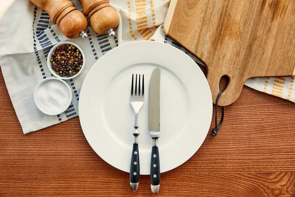 Vista superior de la placa con cuchillo y tenedor cerca de tela con tabla de cortar, cuencos, pimienta y molinos de sal sobre fondo de madera - foto de stock