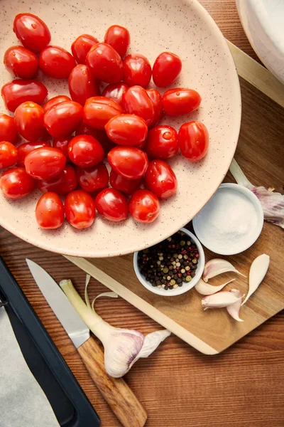 Vista superior del plato con tomates, ajo, cuencos con pimienta y sal sobre tabla de cortar cerca de cuchillo y bandeja de horno sobre fondo de madera - foto de stock