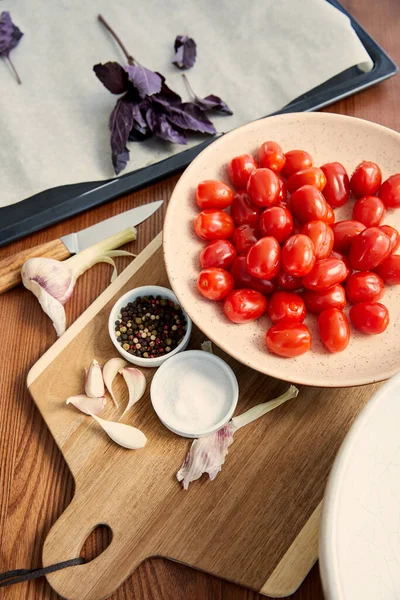 Vista de alto ángulo de la placa con tomates en la tabla de cortar cerca de la bandeja del horno con ingredientes sobre fondo de madera - foto de stock