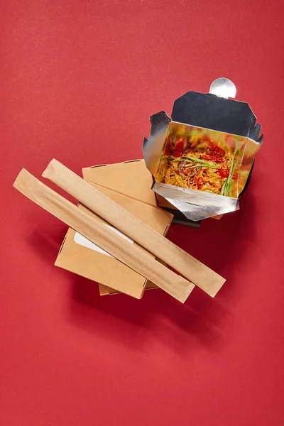 Вид сверху на палочки в бумажной упаковке рядом с острой лапшой в коробке на красном — стоковое фото