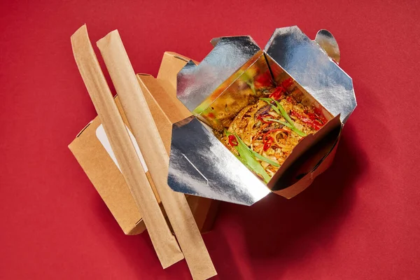 Top view of chopsticks in paper packaging near tasty noodles in takeaway box on red — Stock Photo