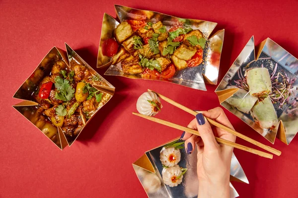 Top view of woman holding chopsticks with steamed bun near tasty chinese food in takeaway boxes on red — Stock Photo